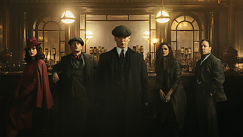 Five people dressed in 1920s-style clothing stand in a dimly lit, elegant bar with vintage decor, glass lamps, and a wooden counter, creating a dramatic and atmospheric scene.