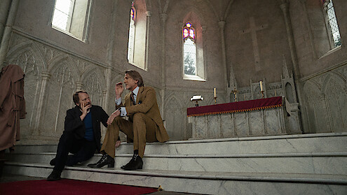 Two men in suits sit on marble steps in a gothic church, talking seriously near an altar with candles and stained glass windows; the setting feels solemn and contemplative.