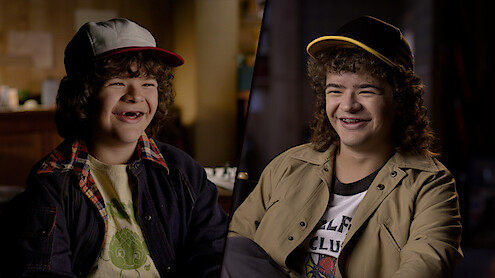 A split-screen image featuring a younger Gaten Matarazzo alongside an older teen version of him, both dressed in casual attire with baseball caps, smiling warmly in softly lit indoor settings.