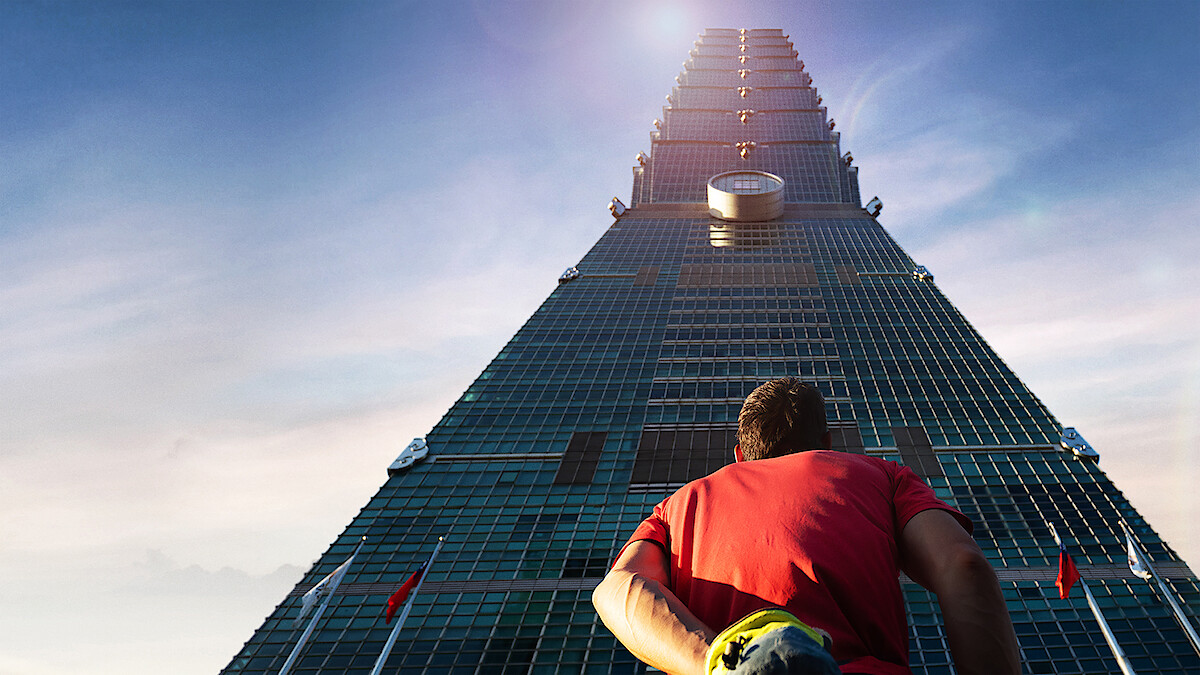Alex Honnold in a red shirt stands at the base of a tall skyscraper, looking up towards the top under a clear blue sky, suggesting ambition or challenge in an urban environment.