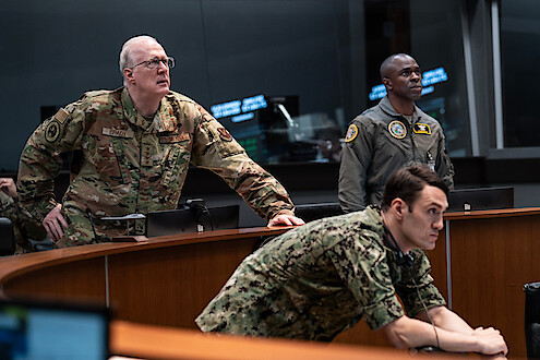 Three military officers in uniform intently observe something off-camera in a modern command center, suggesting a tense and serious atmosphere with strategic or emergency decision-making underway.