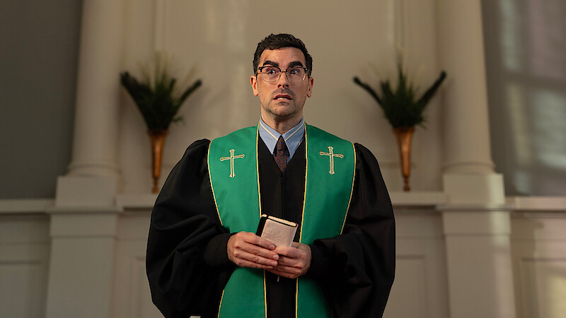 A man dressed in clerical robes with green stole, holding a book, stands in a church sanctuary with white walls, columns, and decorative plants in the background, looking slightly upward.