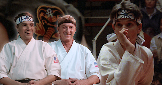 Ralph Macchio and William Zabka sit together in karate uniforms smiling looking at a young Ralph Macchio in 'The Karate Kid.'