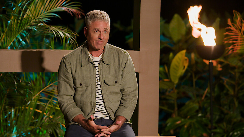 Mark L. Walberg sits outdoors at night, wearing a green jacket and striped shirt, surrounded by tropical plants and a lit torch in the background.