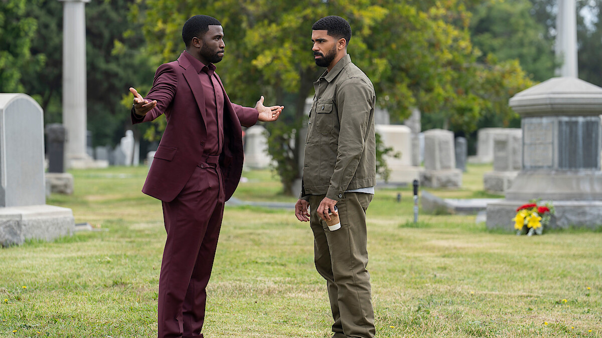 Two men stand in a cemetery, surrounded by gravestones and greenery; one man in a burgundy suit gestures with open arms while the other, in casual clothing, holds a coffee cup and listens.