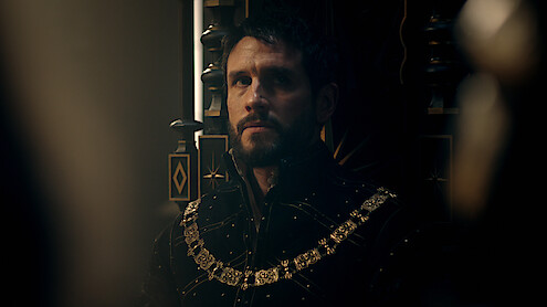 A solemn man in elaborate medieval attire with a gold chain sits on a dark, ornate throne in a dimly lit, regal setting, creating a moody, contemplative atmosphere.
