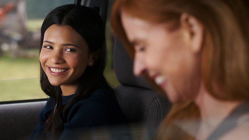Two women sitting in a car, one with dark hair smiling warmly at the other, who has red hair and is laughing. They appear to be enjoying a friendly moment together while parked or driving during the daytime.